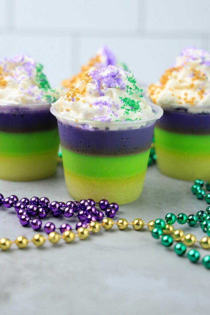 Mardi Gras Jello Shots layered with yellow, green, and purple. On a faux concrete backdrop with Mardi Gras Beads.