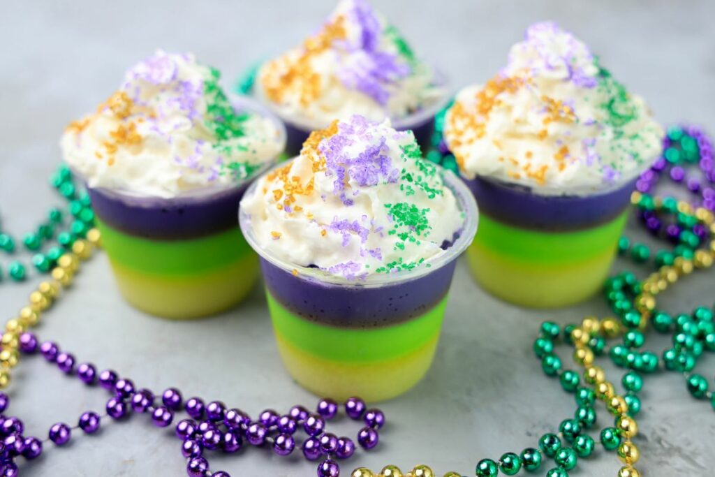 Mardi Gras Jello Shots layered with yellow, green, and purple. On a faux concrete backdrop with Mardi Gras Beads.