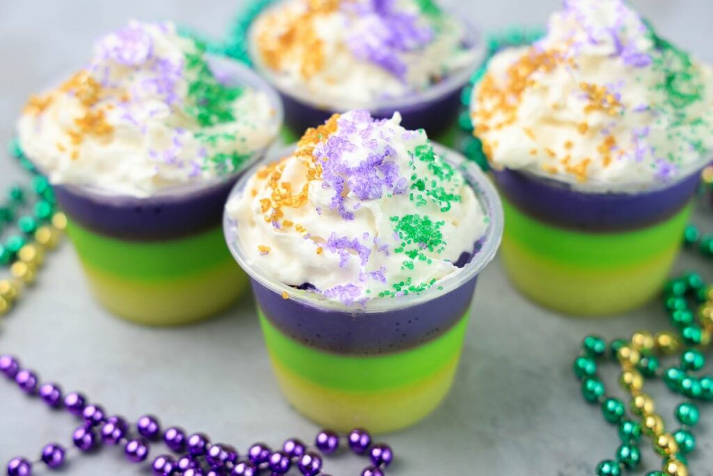 Mardi Gras Jello Shots layered with yellow, green, and purple. On a faux concrete backdrop with Mardi Gras Beads.