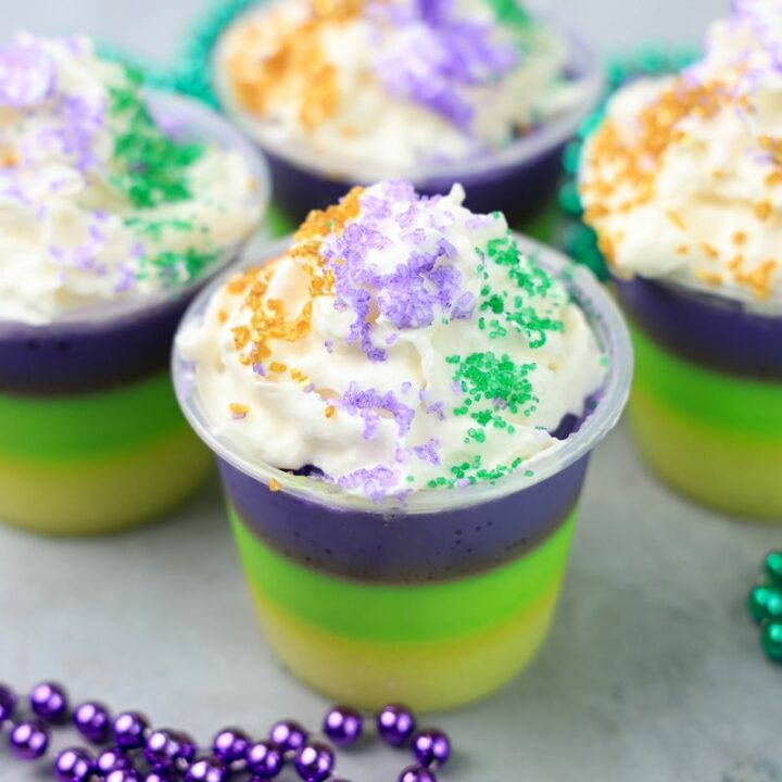 Mardi Gras Jello Shots layered with yellow, green, and purple. On a faux concrete backdrop with Mardi Gras Beads.