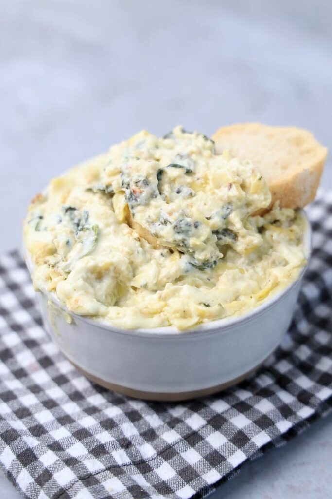 Slow cooker spinach artichoke dip in a white bowl with gray plaid napkin on a faux concrete backdrop.