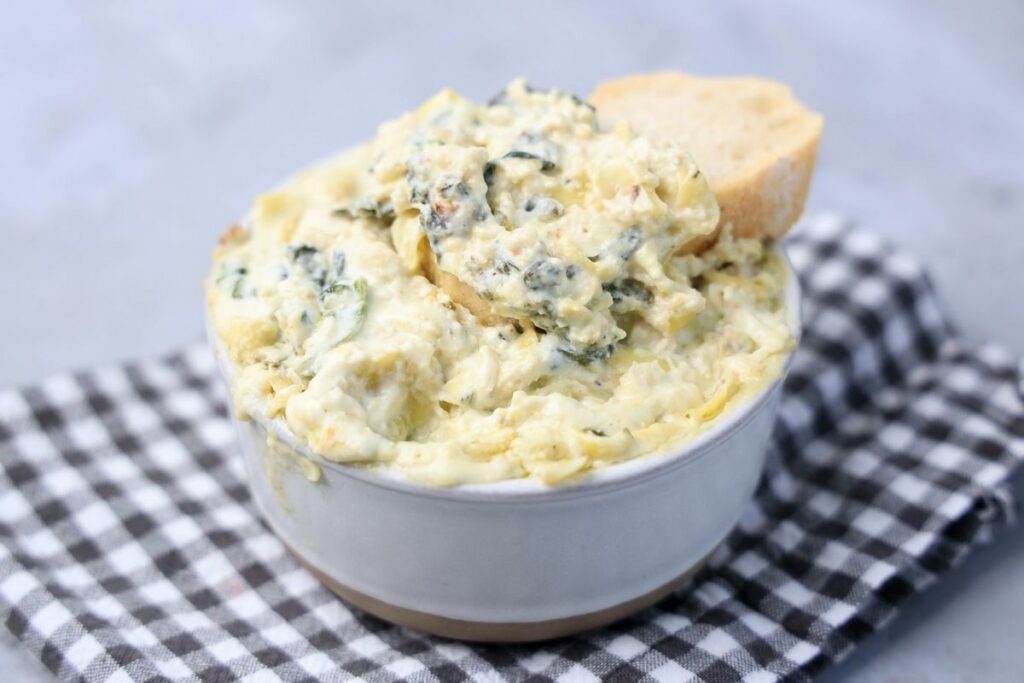 Slow cooker spinach artichoke dip in a white bowl with gray plaid napkin on a faux concrete backdrop.