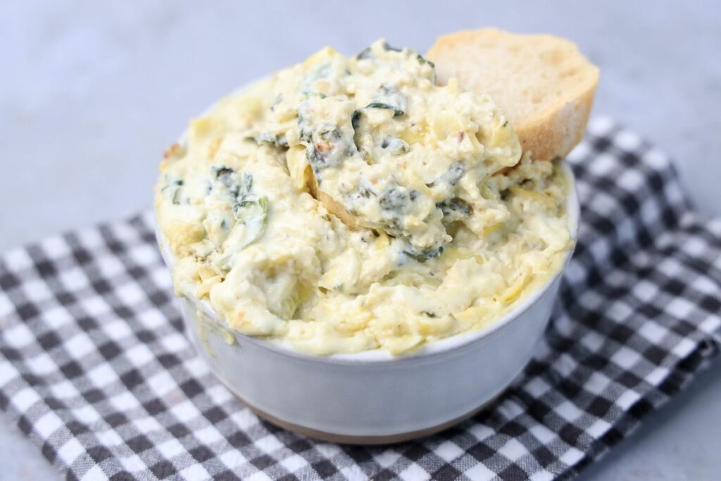 Slow cooker spinach artichoke dip in a white bowl with gray plaid napkin on a faux concrete backdrop.