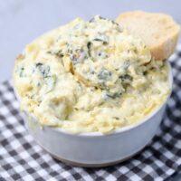 Slow cooker spinach artichoke dip in a white bowl with gray plaid napkin on a faux concrete backdrop.