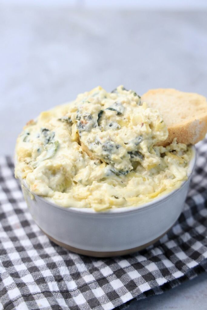 Slow cooker spinach artichoke dip in a white bowl with gray plaid napkin on a faux concrete backdrop.