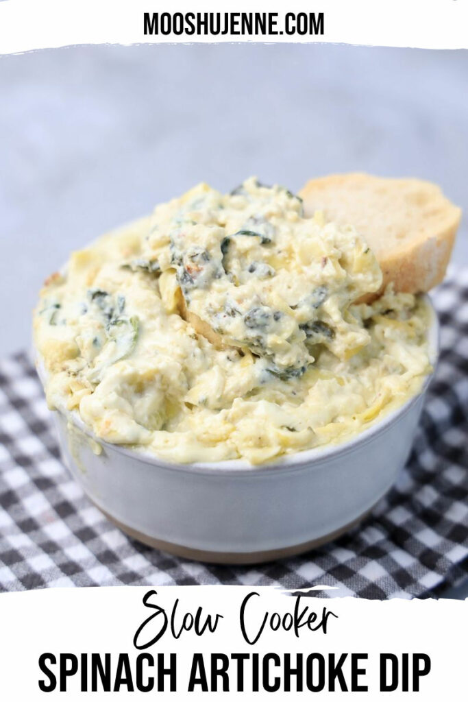 Slow cooker spinach artichoke dip in a white bowl with gray plaid napkin on a faux concrete backdrop.