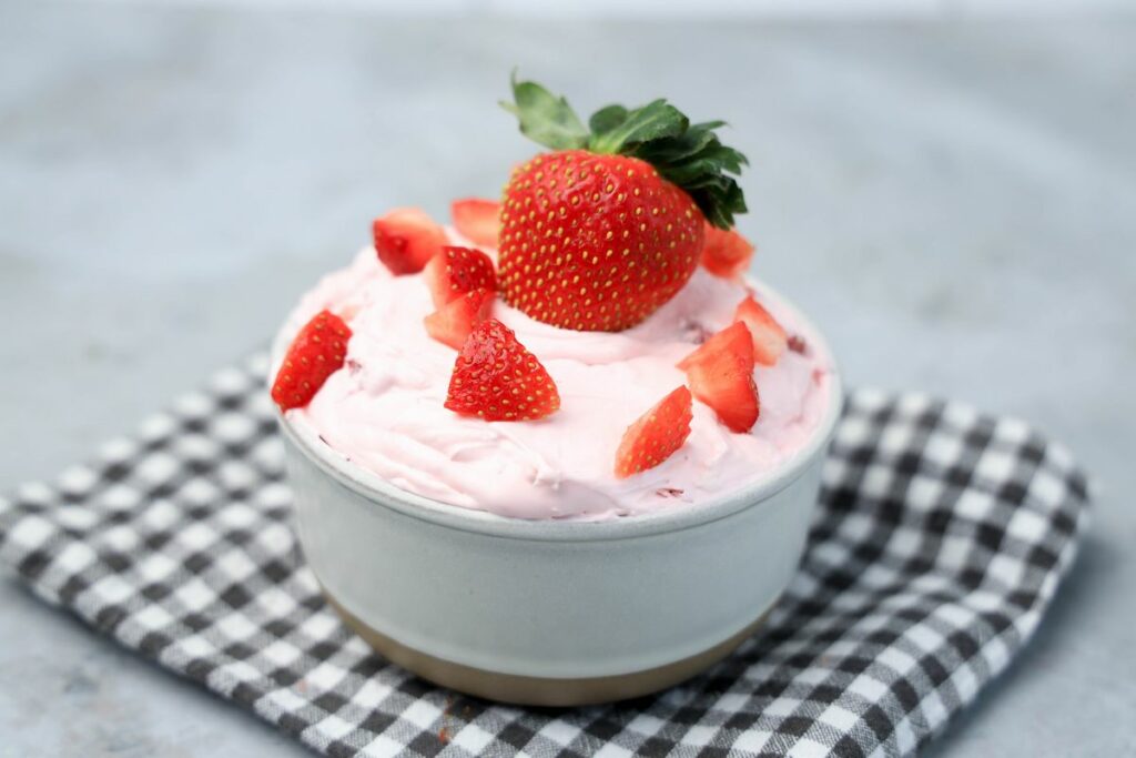 Strawberry cheesecake dip topped with strawberries in a stone bowl with gray plaid napkin on a faux concrete backdrop.