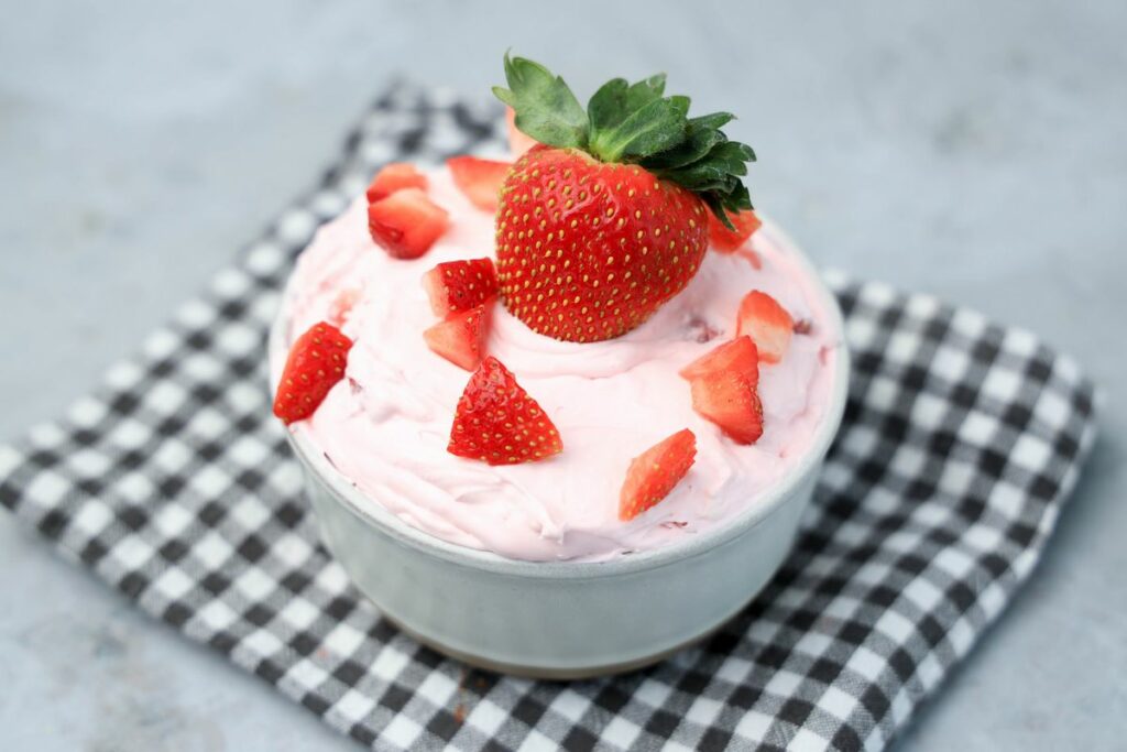 Strawberry cheesecake dip topped with strawberries in a stone bowl with gray plaid napkin on a faux concrete backdrop.