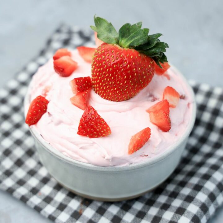 Strawberry cheesecake dip topped with strawberries in a stone bowl with gray plaid napkin on a faux concrete backdrop.