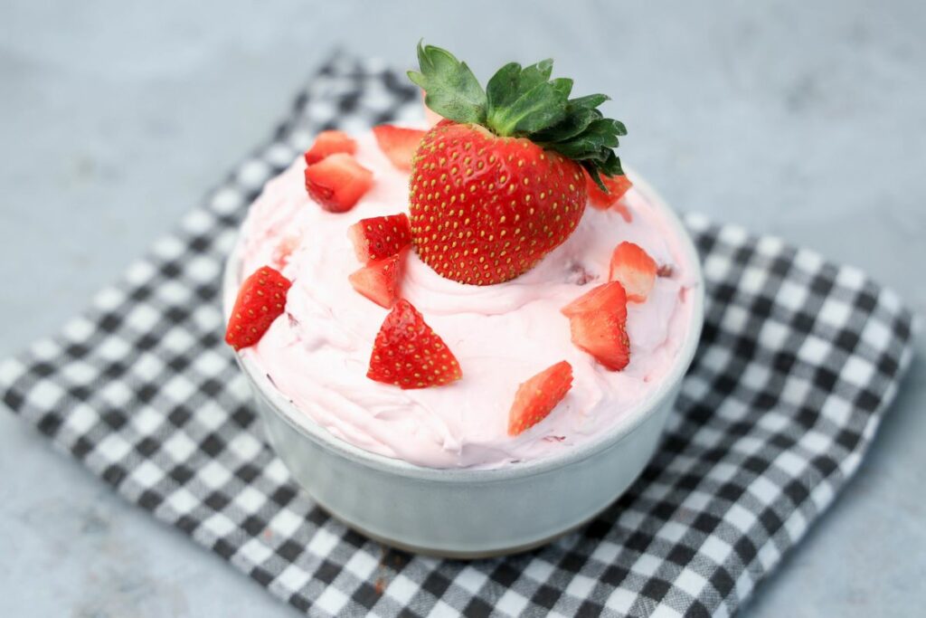 Strawberry cheesecake dip topped with strawberries in a stone bowl with gray plaid napkin on a faux concrete backdrop.