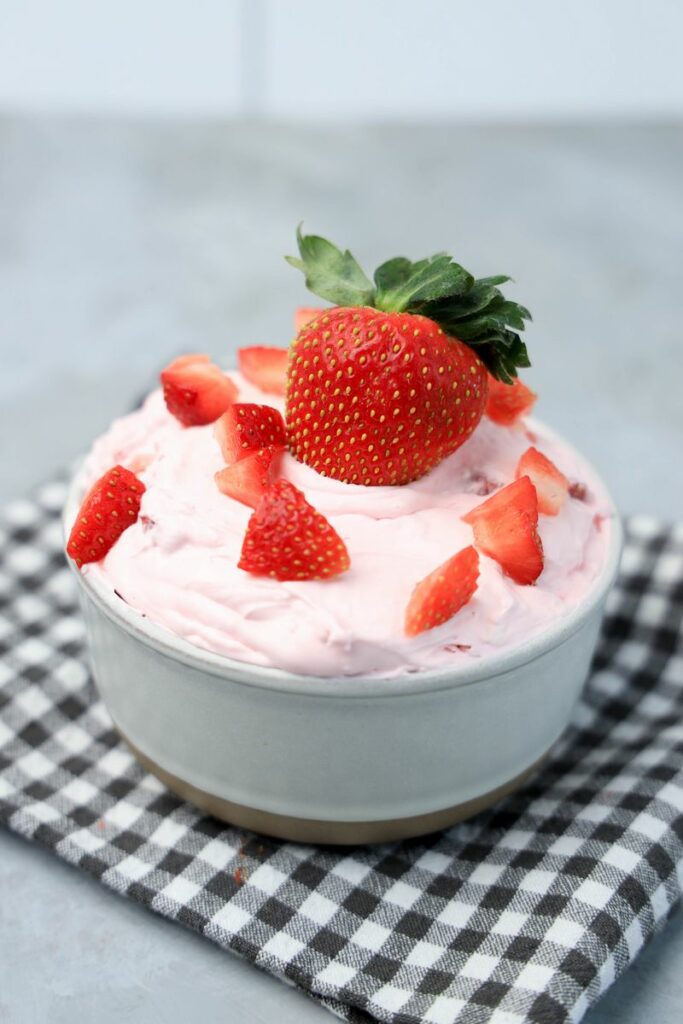 Strawberry cheesecake dip topped with strawberries in a stone bowl with gray plaid napkin on a faux concrete backdrop.