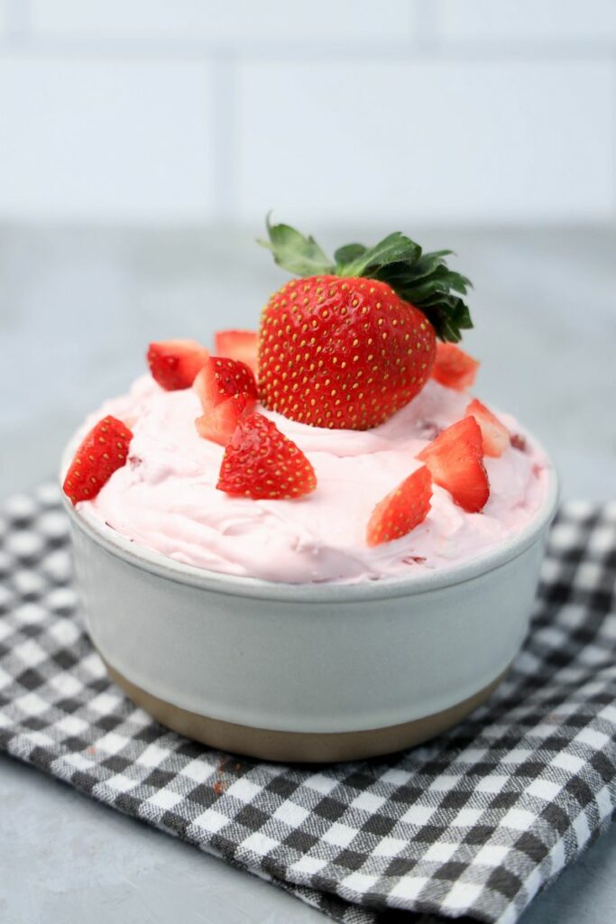 Strawberry cheesecake dip topped with strawberries in a stone bowl with gray plaid napkin on a faux concrete backdrop.
