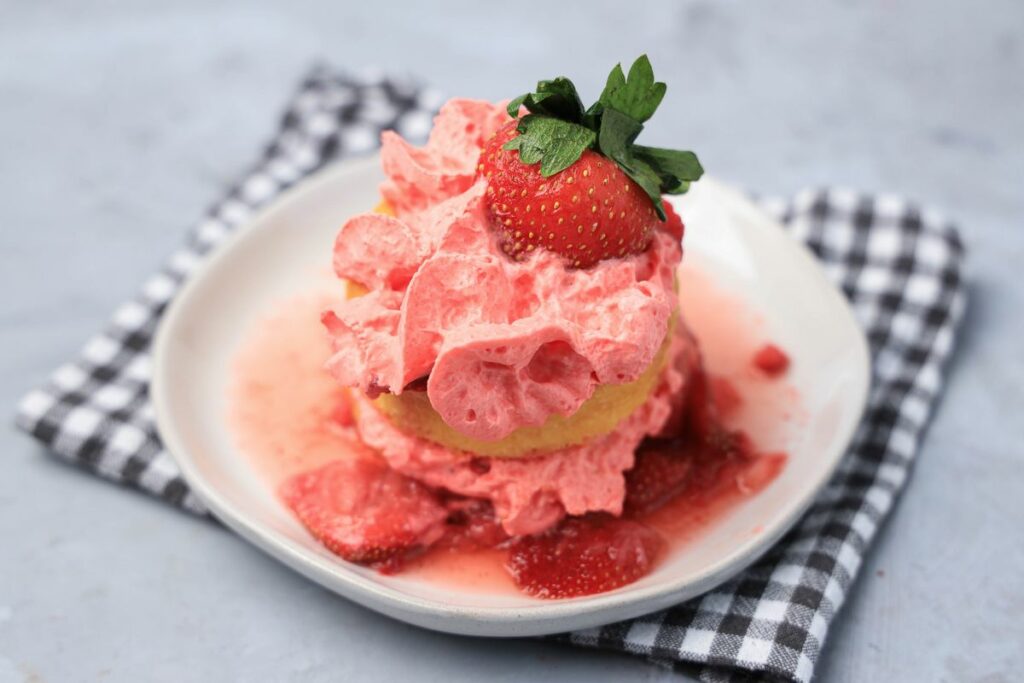 Strawberry shortcake with smashed strawberries and dessert cups with strawberry whipped topping on a white plate with gray plaid napkin.