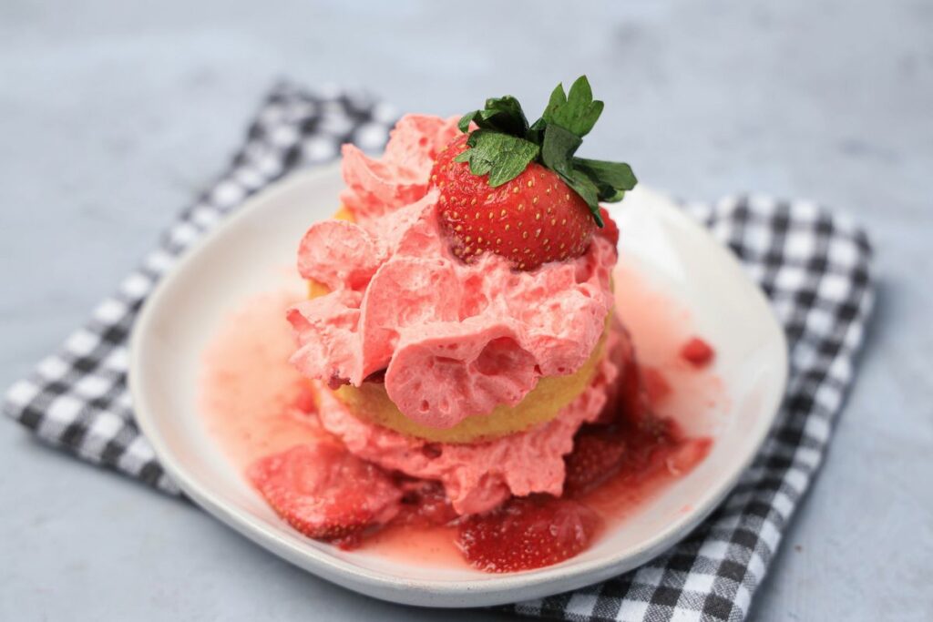 Strawberry shortcake with smashed strawberries and dessert cups with strawberry whipped topping on a white plate with gray plaid napkin.