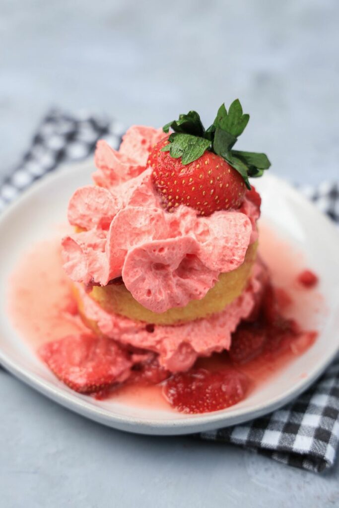 Strawberry shortcake with smashed strawberries and dessert cups with strawberry whipped topping on a white plate with gray plaid napkin.