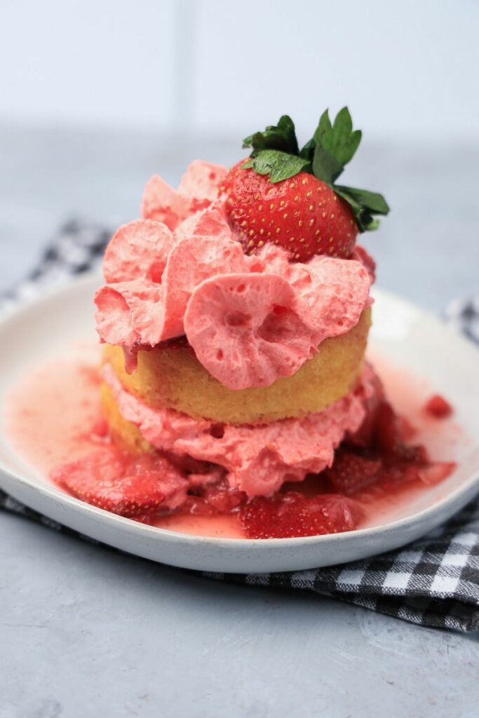 Strawberry shortcake with smashed strawberries and dessert cups with strawberry whipped topping on a white plate with gray plaid napkin.