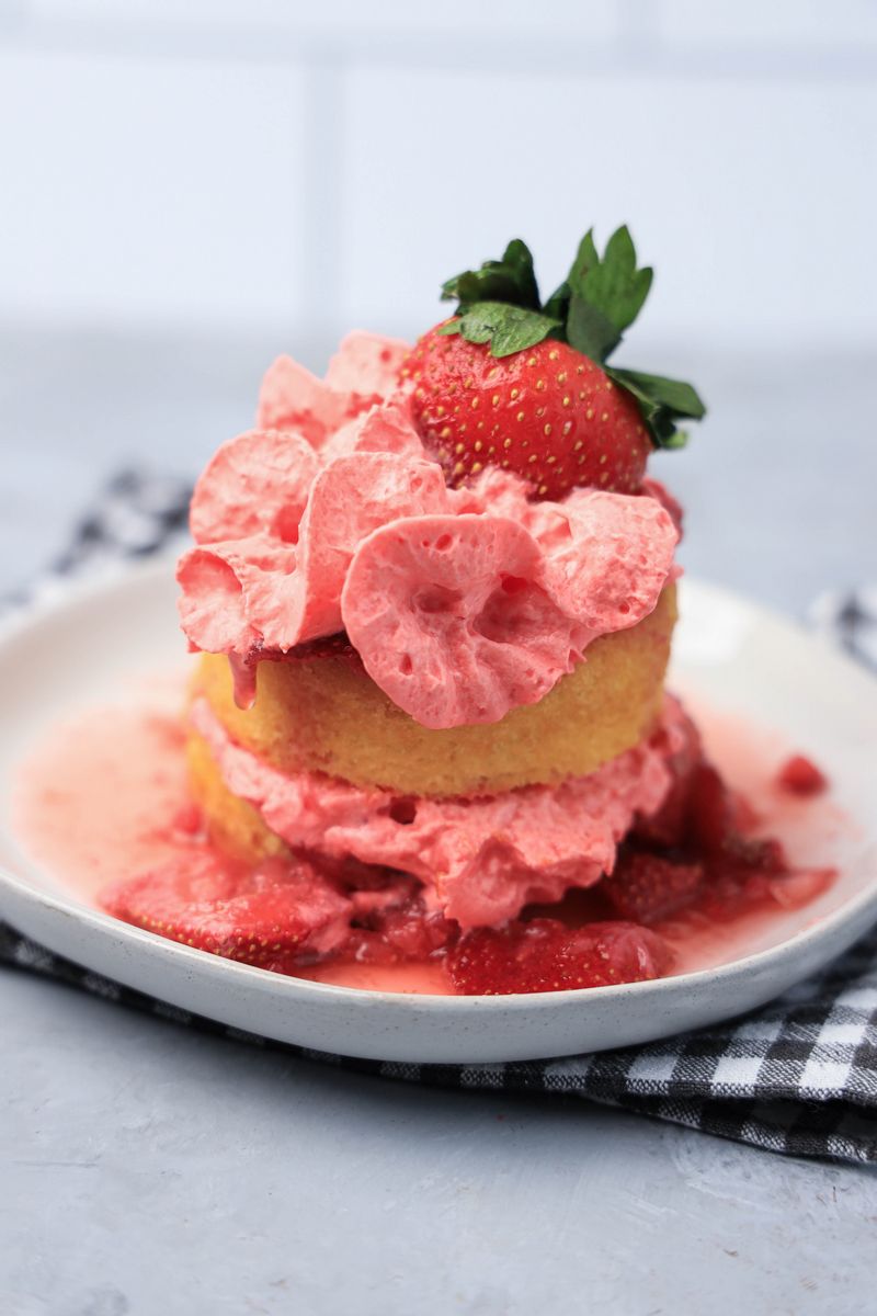 Strawberry shortcake with smashed strawberries and dessert cups with strawberry whipped topping on a white plate with gray plaid napkin.