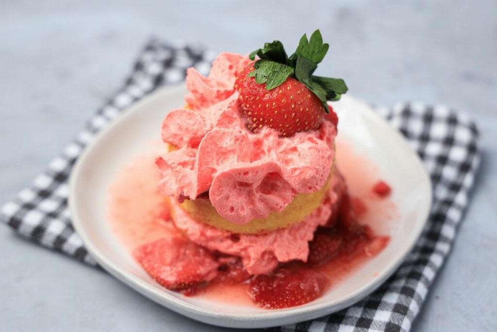 Strawberry shortcake with smashed strawberries and dessert cups with strawberry whipped topping on a white plate with gray plaid napkin.