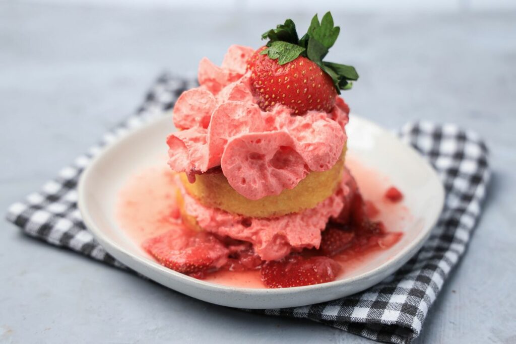 Strawberry shortcake with smashed strawberries and dessert cups with strawberry whipped topping on a white plate with gray plaid napkin.