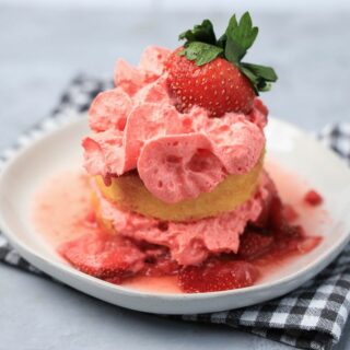 Strawberry shortcake with smashed strawberries and dessert cups with strawberry whipped topping on a white plate with gray plaid napkin.