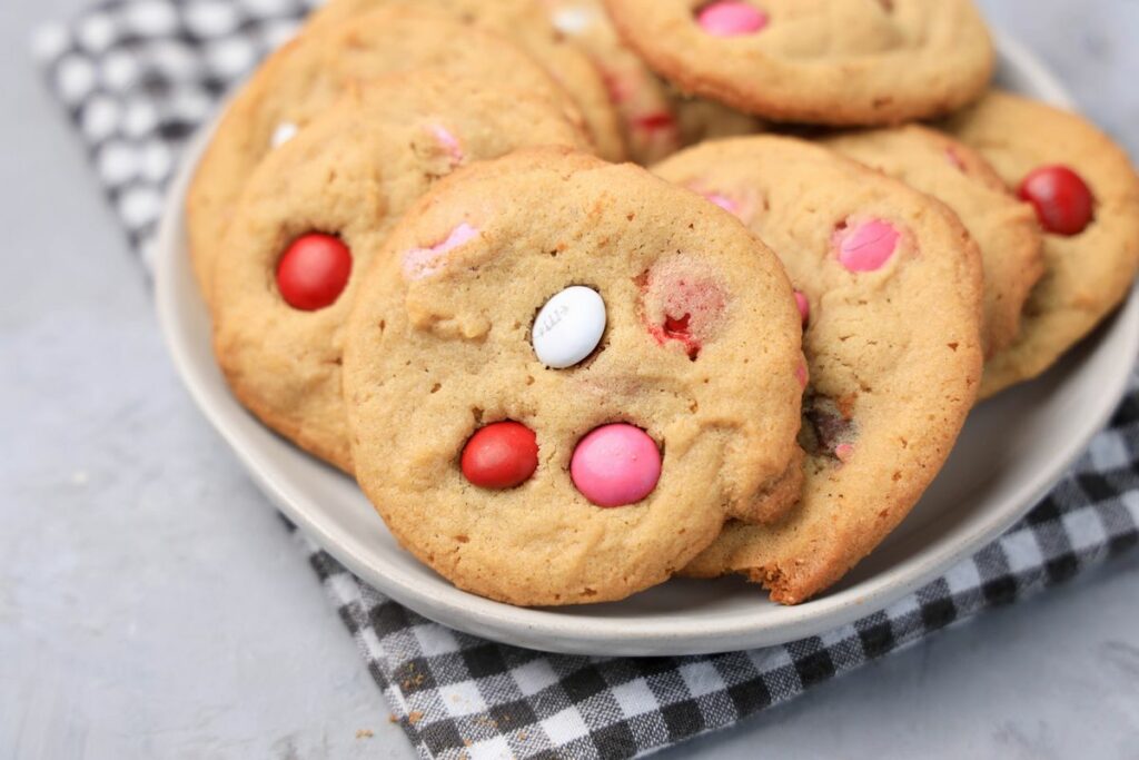 Valentine's Peanut Butter M&M cookies on a white plate with gray plaid napkin on a faux concrete backdrop.