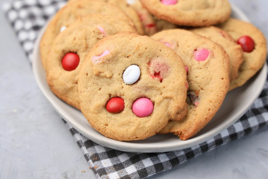 Valentine's Peanut Butter M&M cookies on a white plate with gray plaid napkin on a faux concrete backdrop.