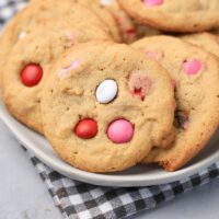 Valentine's Peanut Butter M&M cookies on a white plate with gray plaid napkin on a faux concrete backdrop.