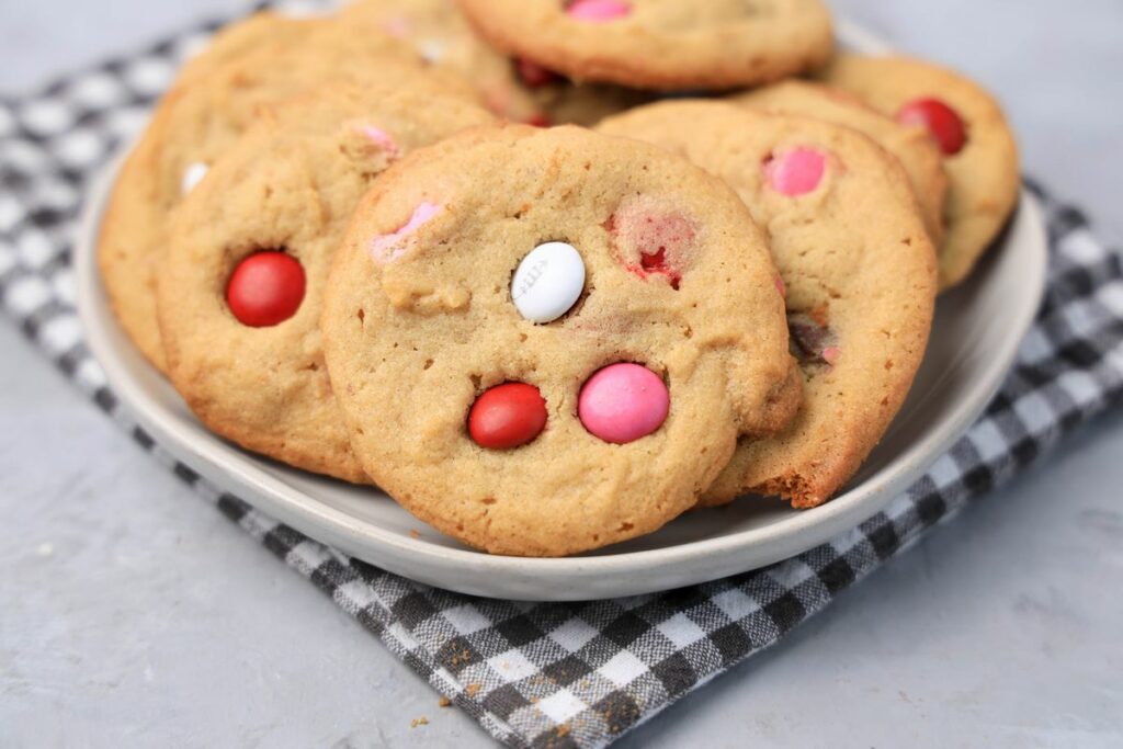 Valentine's Peanut Butter M&M cookies on a white plate with gray plaid napkin on a faux concrete backdrop.