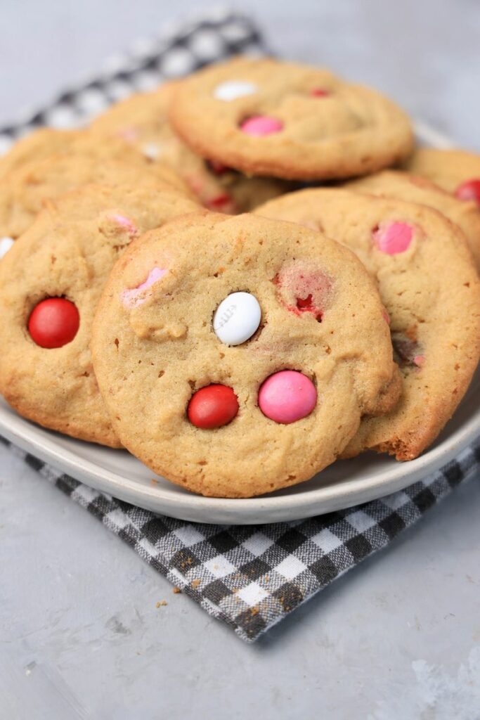 Valentine's Peanut Butter M&M cookies on a white plate with gray plaid napkin on a faux concrete backdrop.