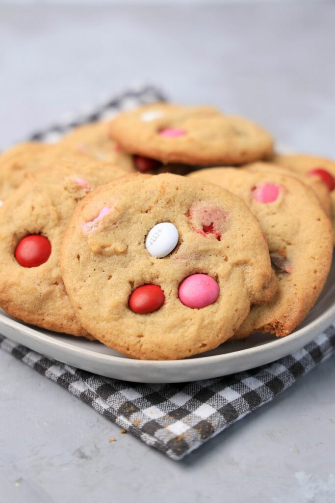 Valentine's Peanut Butter M&M cookies on a white plate with gray plaid napkin on a faux concrete backdrop.