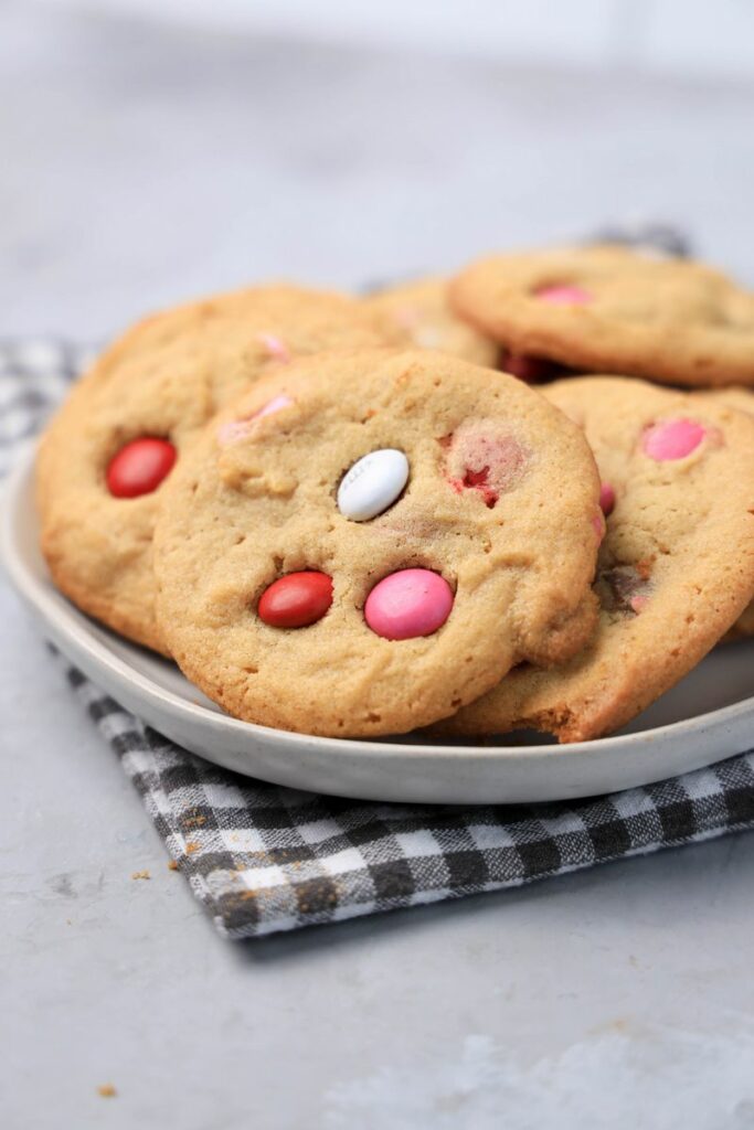 Valentine's Peanut Butter M&M cookies on a white plate with gray plaid napkin on a faux concrete backdrop.