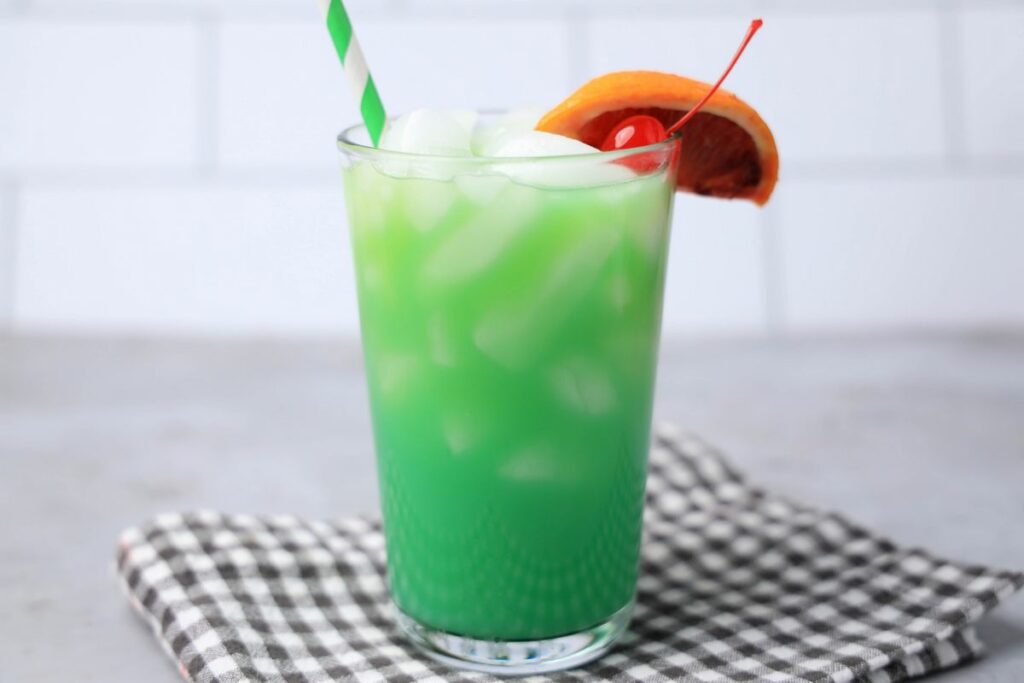 Drunken Leprechaun cocktail with a green striped straw on a gray plaid napkin with a faux concrete backdrop.