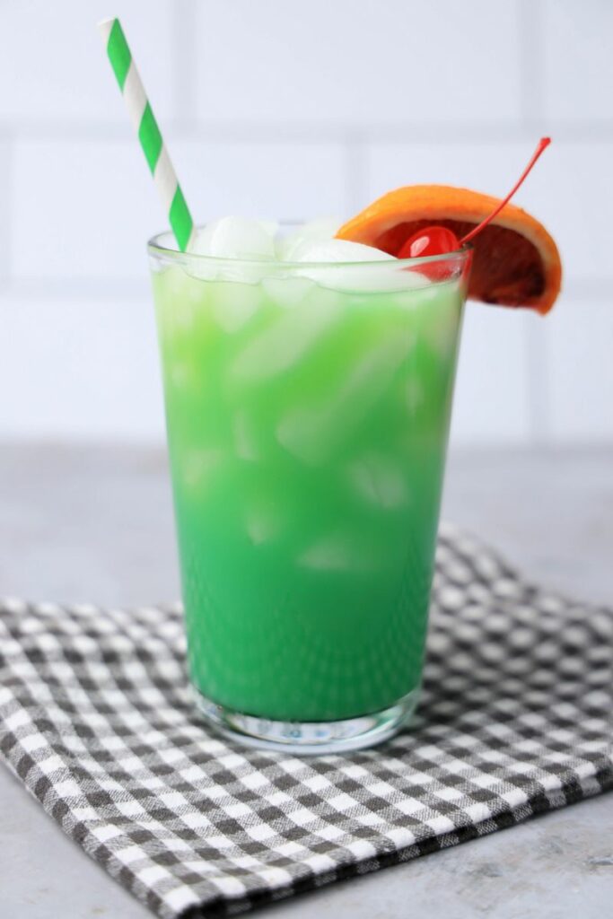 Drunken Leprechaun cocktail with a green striped straw on a gray plaid napkin with a faux concrete backdrop.