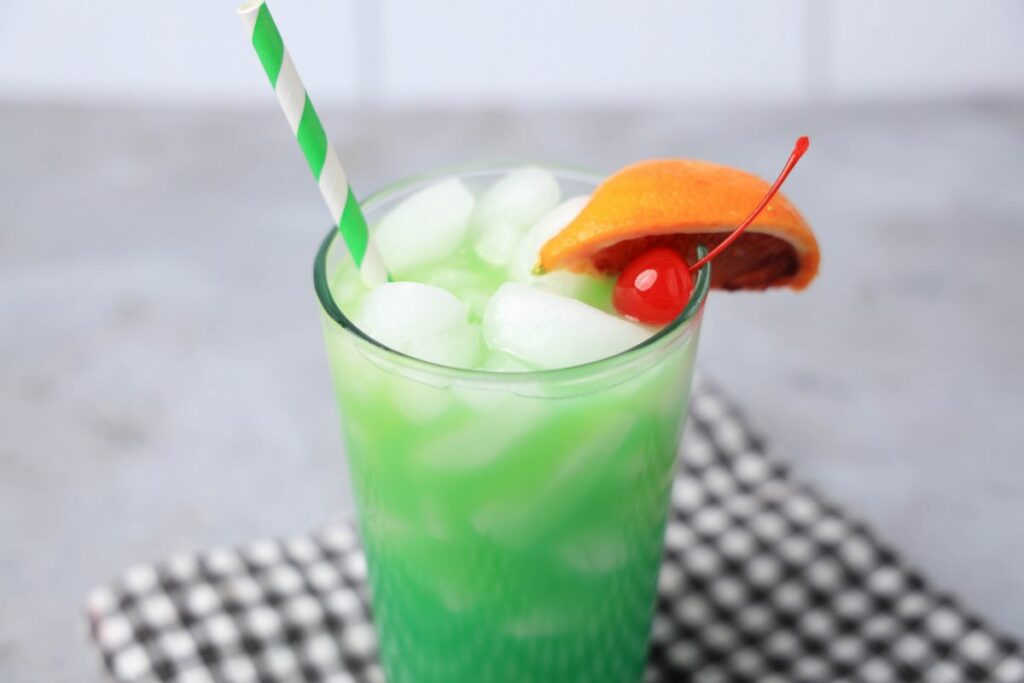 Drunken Leprechaun cocktail with a green striped straw on a gray plaid napkin with a faux concrete backdrop.