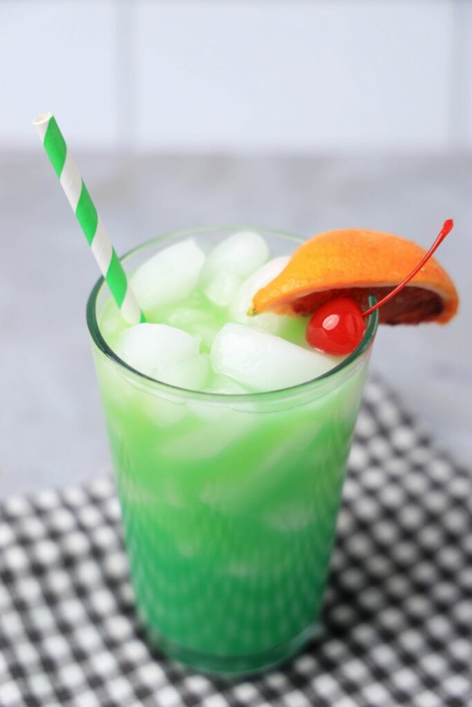 Drunken Leprechaun cocktail with a green striped straw on a gray plaid napkin with a faux concrete backdrop.