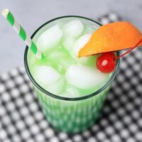 Drunken Leprechaun cocktail with a green striped straw on a gray plaid napkin with a faux concrete backdrop.