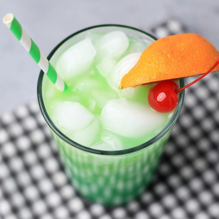 Drunken Leprechaun cocktail with a green striped straw on a gray plaid napkin with a faux concrete backdrop.