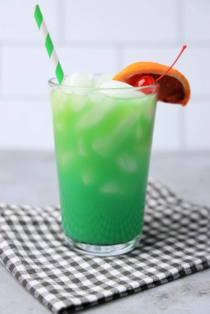 Drunken Leprechaun cocktail with a green striped straw on a gray plaid napkin with a faux concrete backdrop.