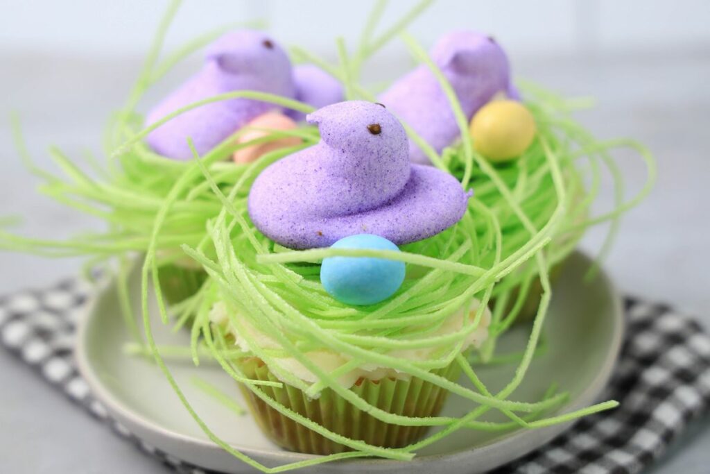 Peeps Chick Nest Cupcakes with green edible grass on a white plate with  a gray plaid napkin on a faux concrete backdrop.