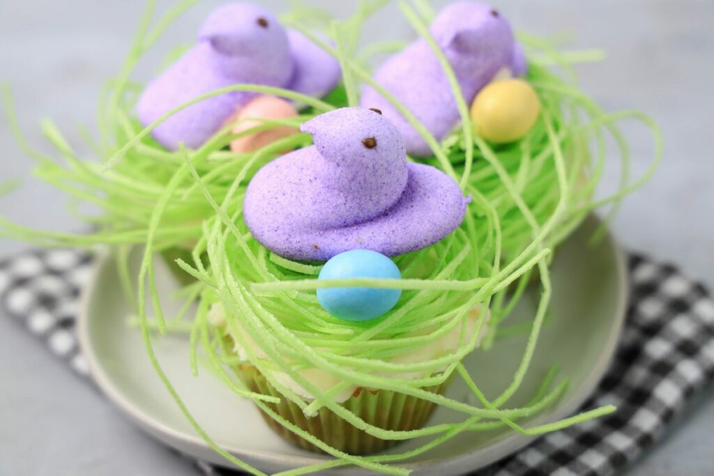 Peeps Chick Nest Cupcakes with green edible grass on a white plate with a gray plaid napkin on a faux concrete backdrop.