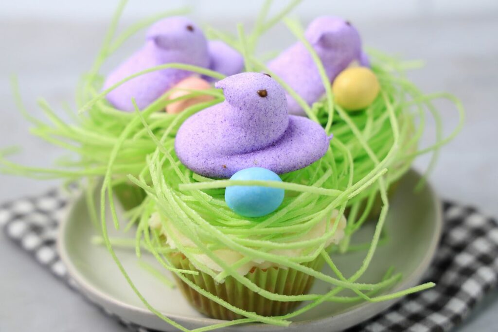 Peeps Chick Nest Cupcakes with green edible grass on a white plate with a gray plaid napkin on a faux concrete backdrop.