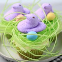 Peeps Chick Nest Cupcakes with green edible grass on a white plate with a gray plaid napkin on a faux concrete backdrop.