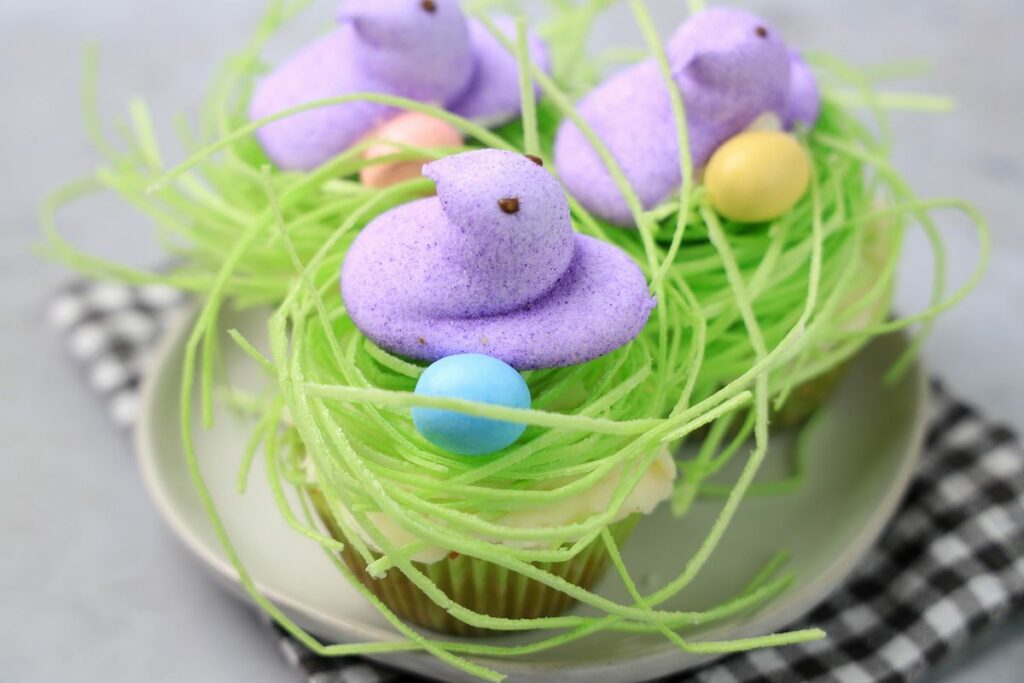 Peeps Chick Nest Cupcakes with green edible grass on a white plate with a gray plaid napkin on a faux concrete backdrop.