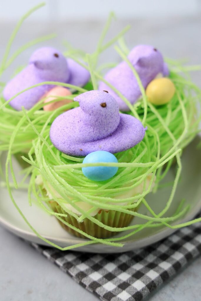 Peeps Chick Nest Cupcakes with green edible grass on a white plate with a gray plaid napkin on a faux concrete backdrop.