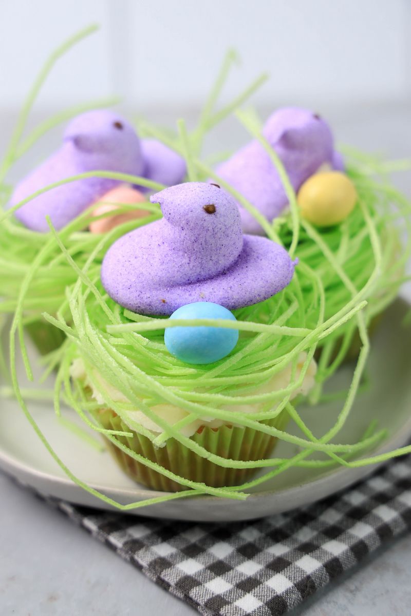 Peeps Chick Nest Cupcakes with green edible grass on a white plate with a gray plaid napkin on a faux concrete backdrop.