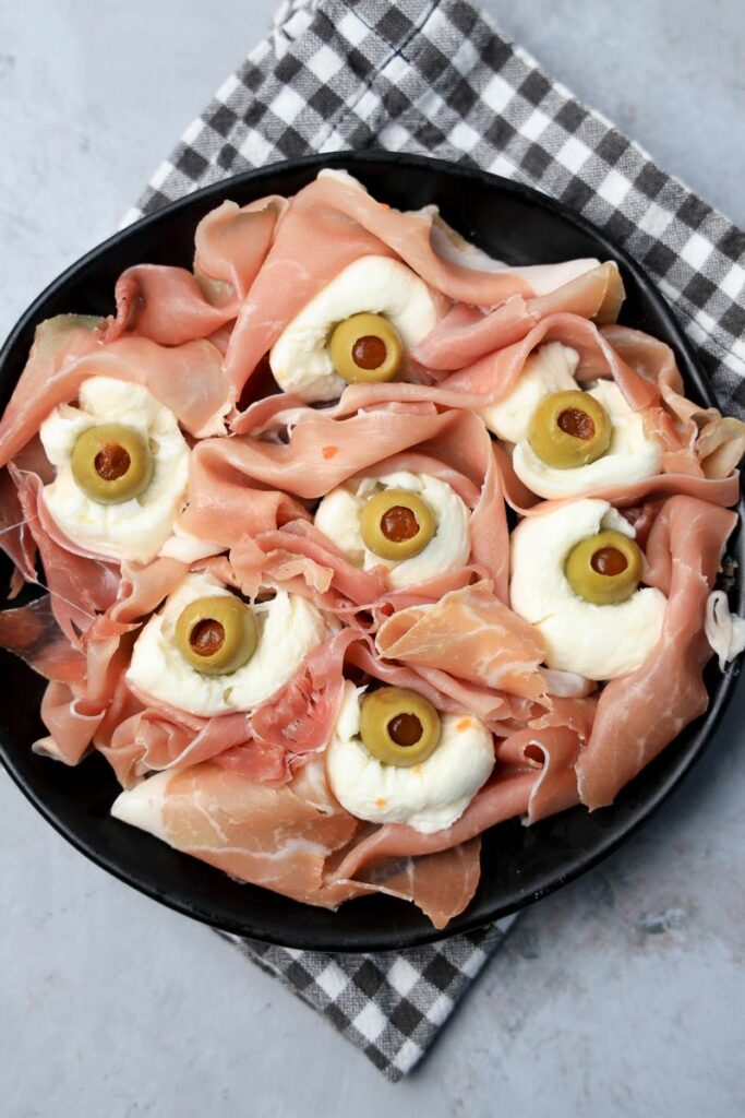 Fresh mozzarella, green olives, and prosciutto to make spooky eyeballs on a black plate.