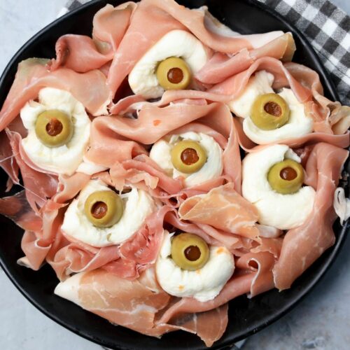 Fresh mozzarella, green olives, and prosciutto to make spooky eyeballs on a black plate.