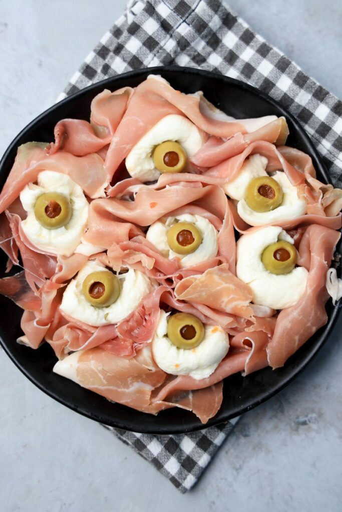 Fresh mozzarella, green olives, and prosciutto to make spooky eyeballs on a black plate.