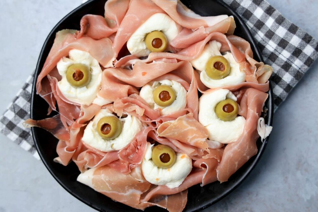 Fresh mozzarella, green olives, and prosciutto to make spooky eyeballs on a black plate.