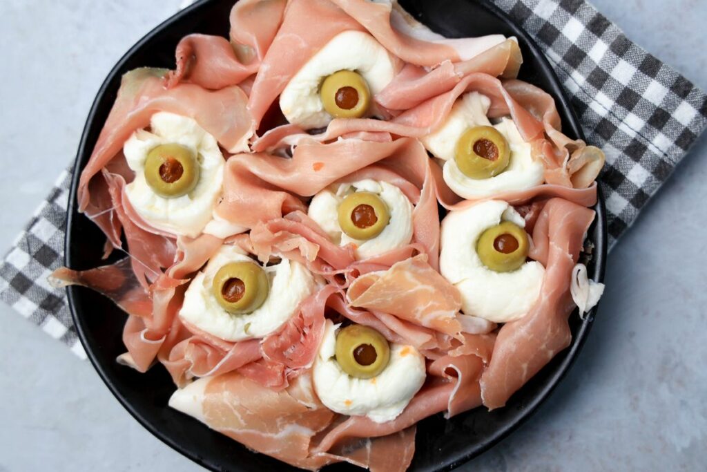 Fresh mozzarella, green olives, and prosciutto to make spooky eyeballs on a black plate.